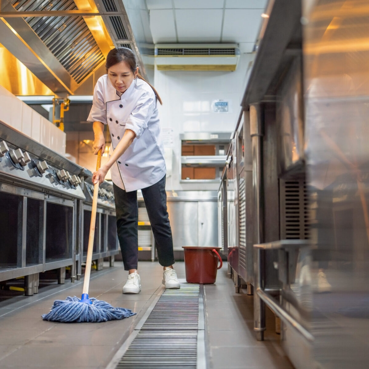 A woman with dark hair in a commercial kitchen. The fixtures are all shiny, steel and she is wearing a chef jacket. She is mopping the floor