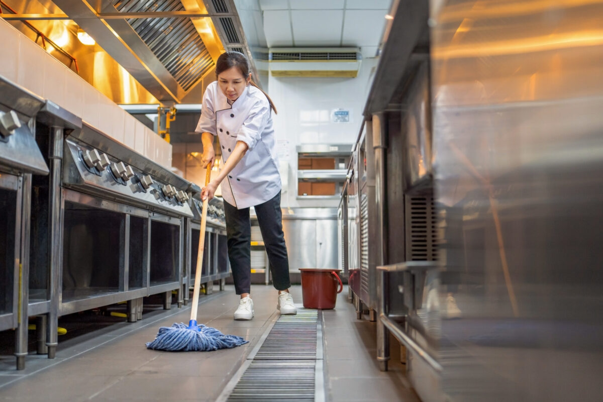 A woman with dark hair in a commercial kitchen. The fixtures are all shiny, steel and she is wearing a chef jacket. She is mopping the floor