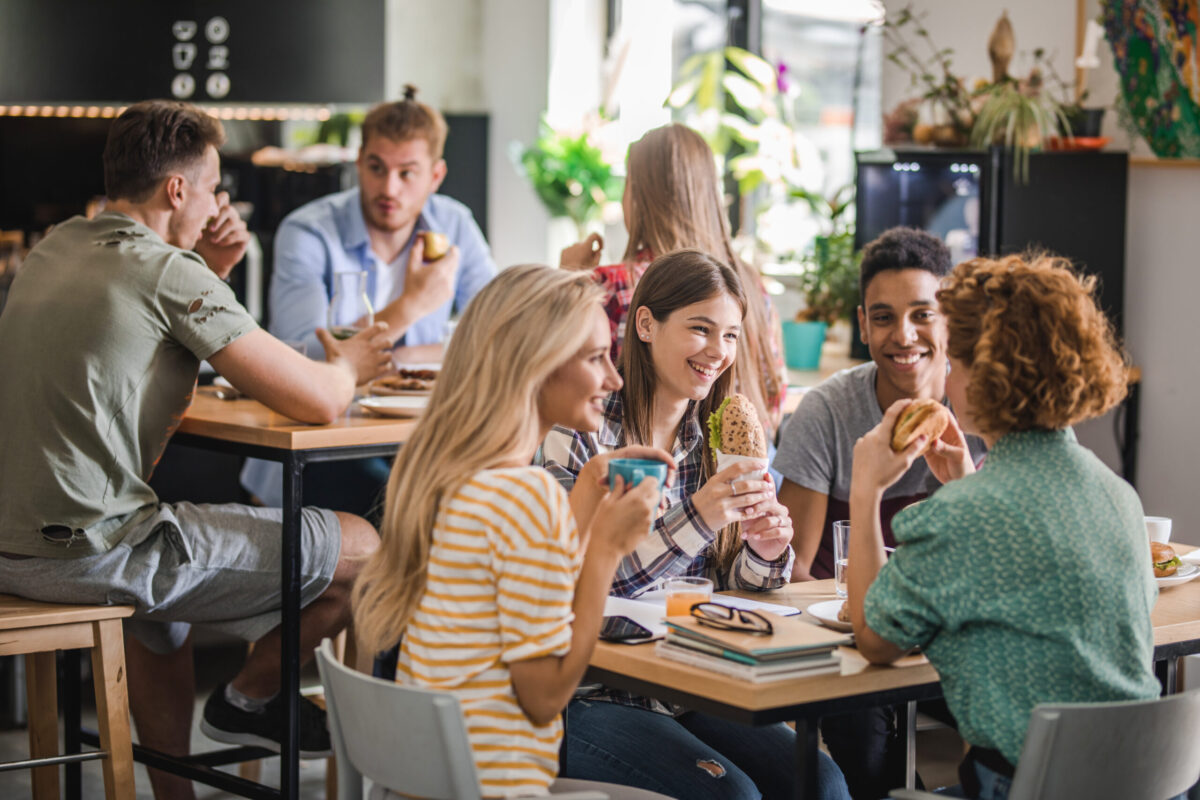 University students sat at tables in a cafe area. Eating lunch and smiling