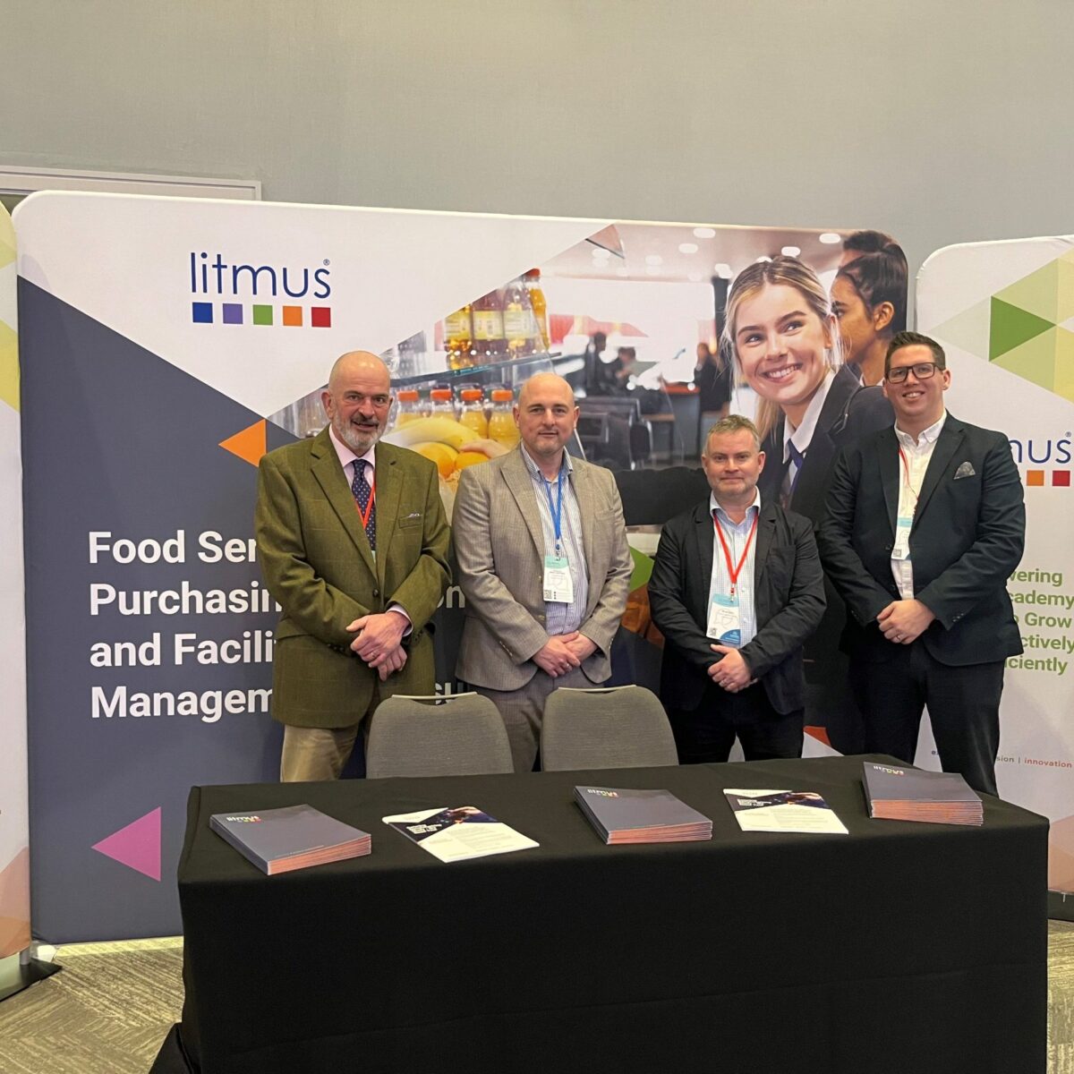 The Litmus team at the Confederation of School Trusts (CST) Finance and Operations Conference 2025. The team are stoofd in front of the Litmus stand. There are 4 of the team, all wearing suits.