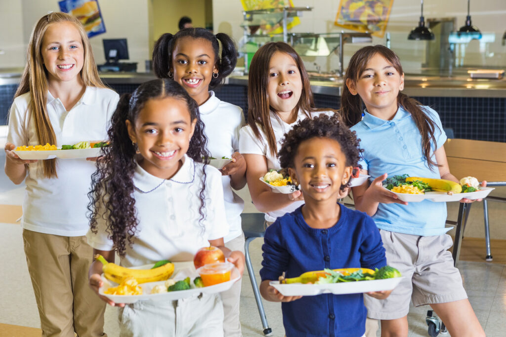 6 school children holding lunch trays filled with healthy food. The children are smiling and happy