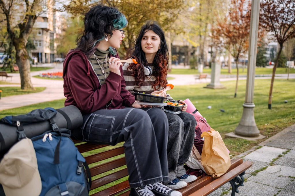 2 gen alpha girls sat on the back of a bench in a park/. Both girls are eating lunch. They are dress in a grunge style with jeans and dyed hair.