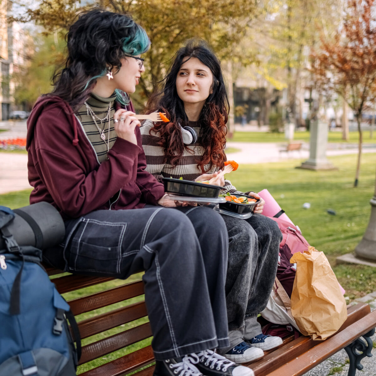2 gen alpha girls sat on the back of a bench in a park/. Both girls are eating lunch. They are dress in a grunge style with jeans and dyed hair.