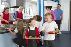Primary school children collecting their lunches from the counter on trays. There is a boy and a girl and they are smiling at each other.