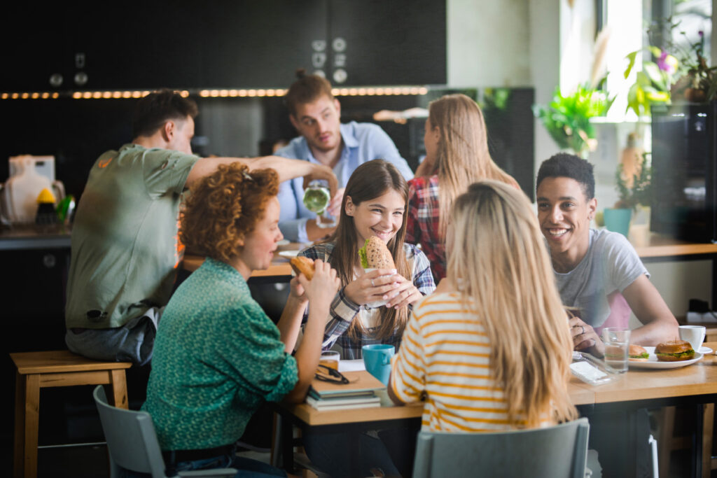 A group of university students sat at a table in the canteen eating sandwiches and smiling