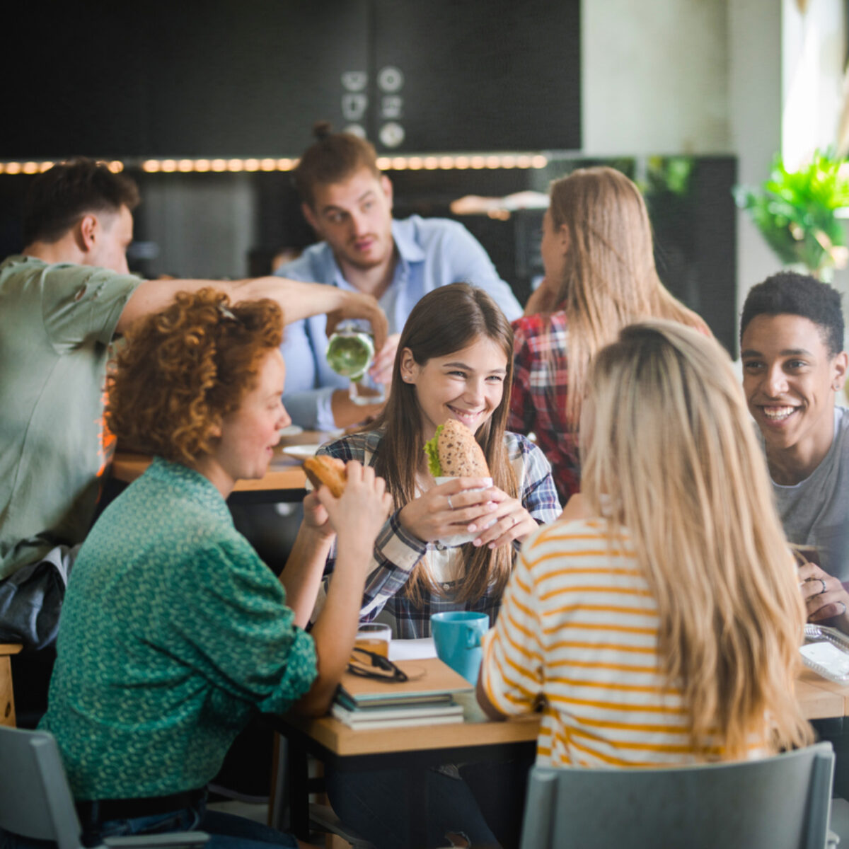A group of university students sat at a table in the canteen eating sandwiches and smiling