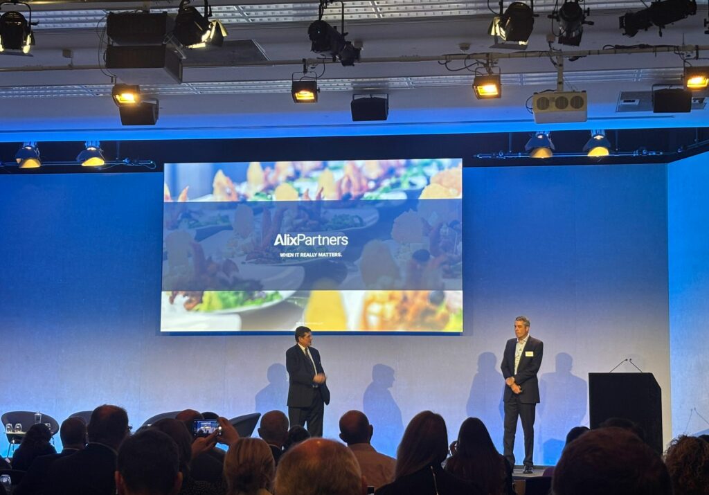 The stage at the Alix Partners event as seen from the audience. There are 2 presenters and a large screen